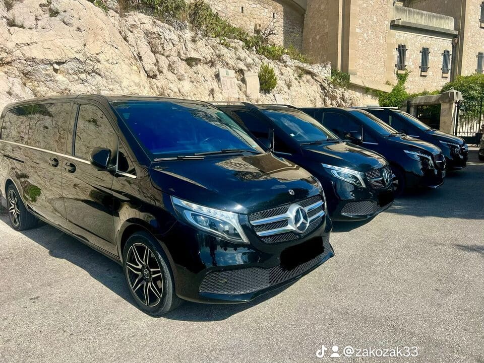 Plusieurs Mercedes-Benz Vito noirs garés en ligne dans un stationnement extérieur, devant un bâtiment en pierre