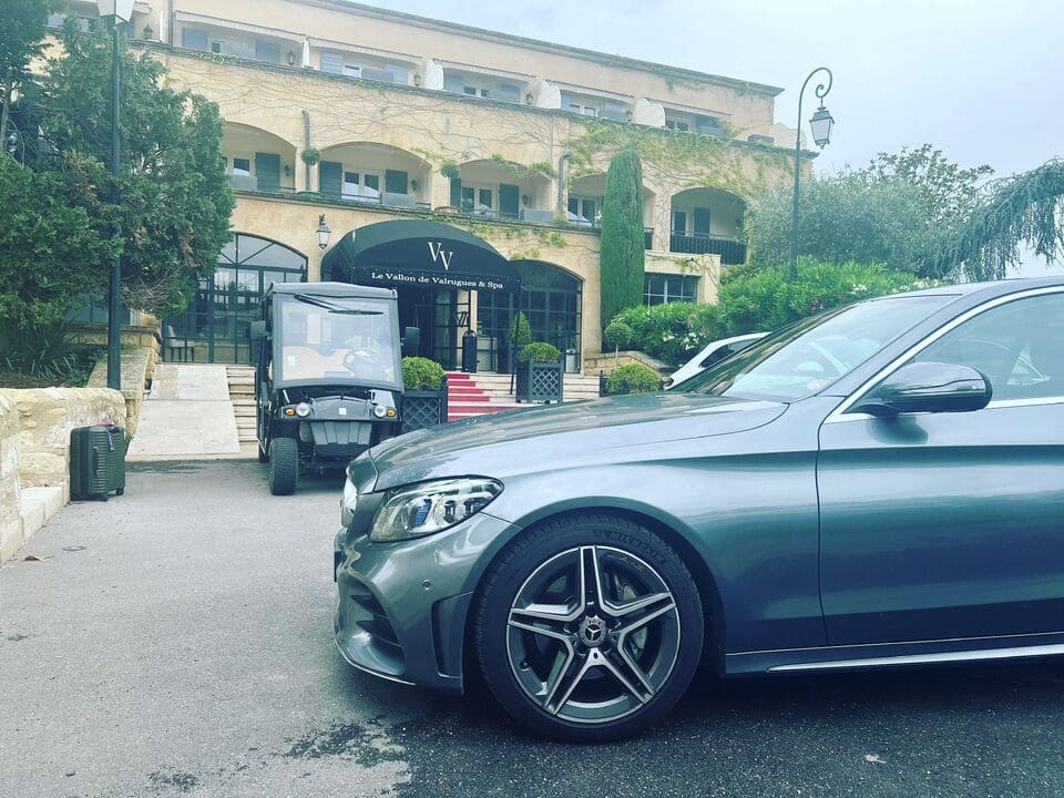 Voiture de luxe grise garée devant un hôtel avec un chariot de golf et une valise à côté de l'entrée.