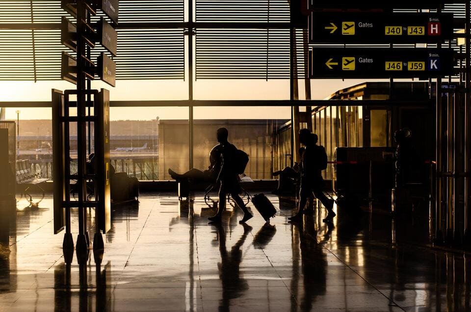 Silhouette de voyageurs tirant leurs valises dans un aéroport au coucher du soleil, avec des panneaux indicateurs de portes au-dessus.