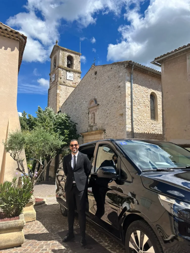 Un chauffeur en costume noir près d'un van noir, avec une église et son clocher en arrière-plan dans un village.