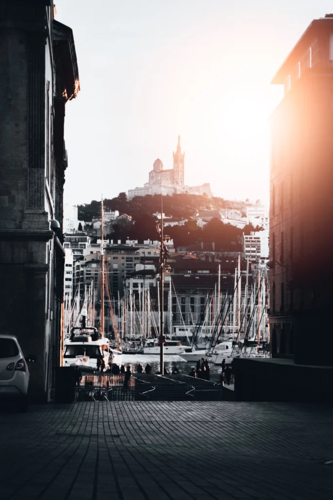 Vue du vieux port de Marseille avec de nombreux bateaux amarrés et la basilique Notre-Dame de la Garde perchée sur la colline au loin.