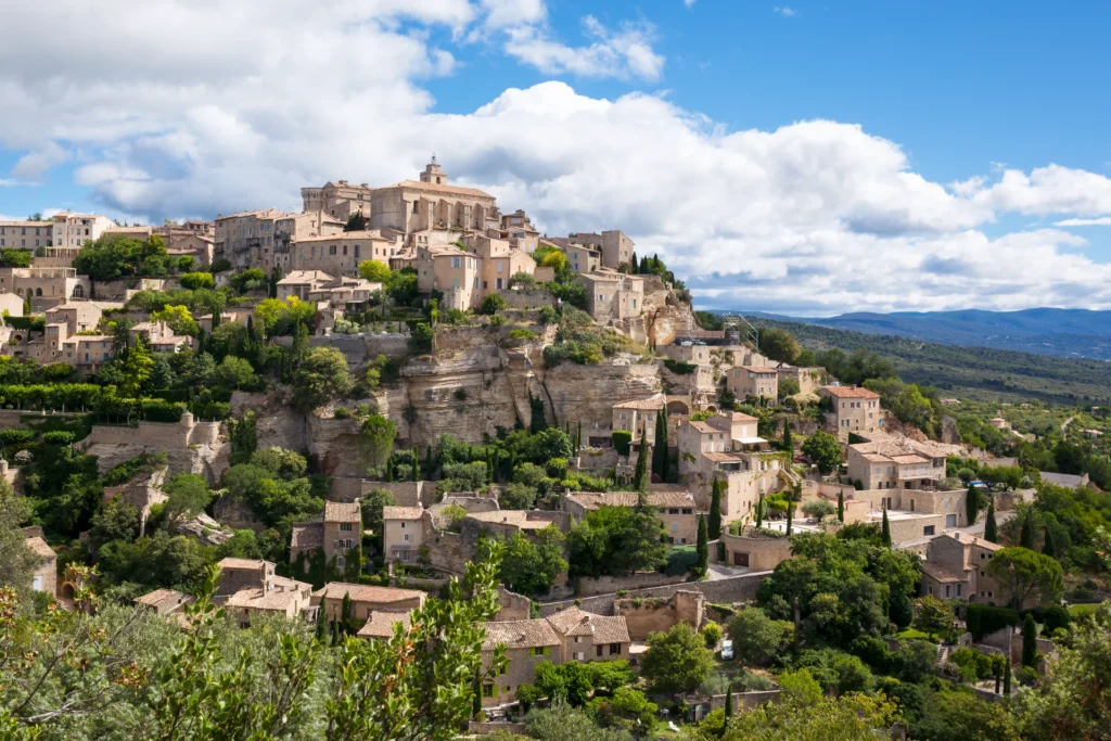 Vue d'ensemble du village perché de Gordes, en Provence, avec ses maisons en pierre typiques dominant une colline verdoyante sous un ciel bleu. Chauffeur VTC Circuit Touristique Alpilles