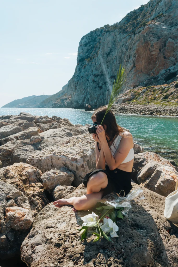 Une jeune femme assise sur des rochers au bord de la mer, prenant une photo avec un appareil photo, avec des fleurs blanches posées à côté d'elle.