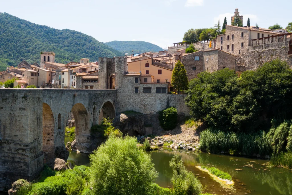 Pont de pierre médiéval avec des arches, traversant une rivière verte, menant à un village pittoresque aux toits oranges et maisons en pierre.