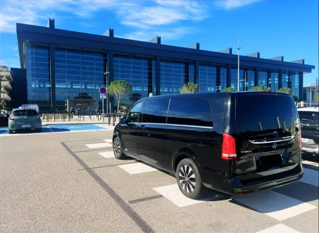 Mercedes Classe V noire garée devant un bâtiment moderne avec de grandes fenêtres bleues, probable aéroport ou gare.
